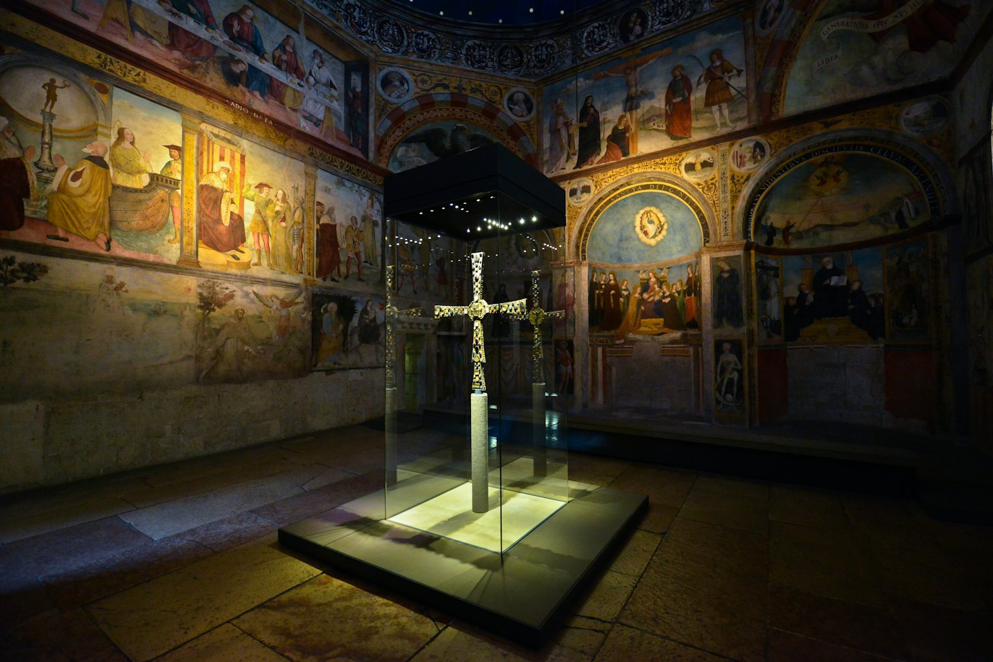 Ornate reliquary cross in a glass case before frescoes.